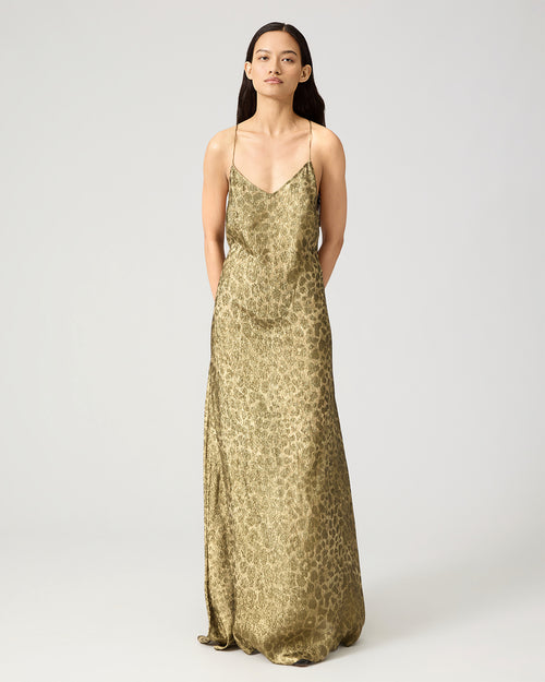 A woman modeling a floor-length satin slip dress with a gold and brown leopard print pattern, photographed against a neutral light gray studio background.