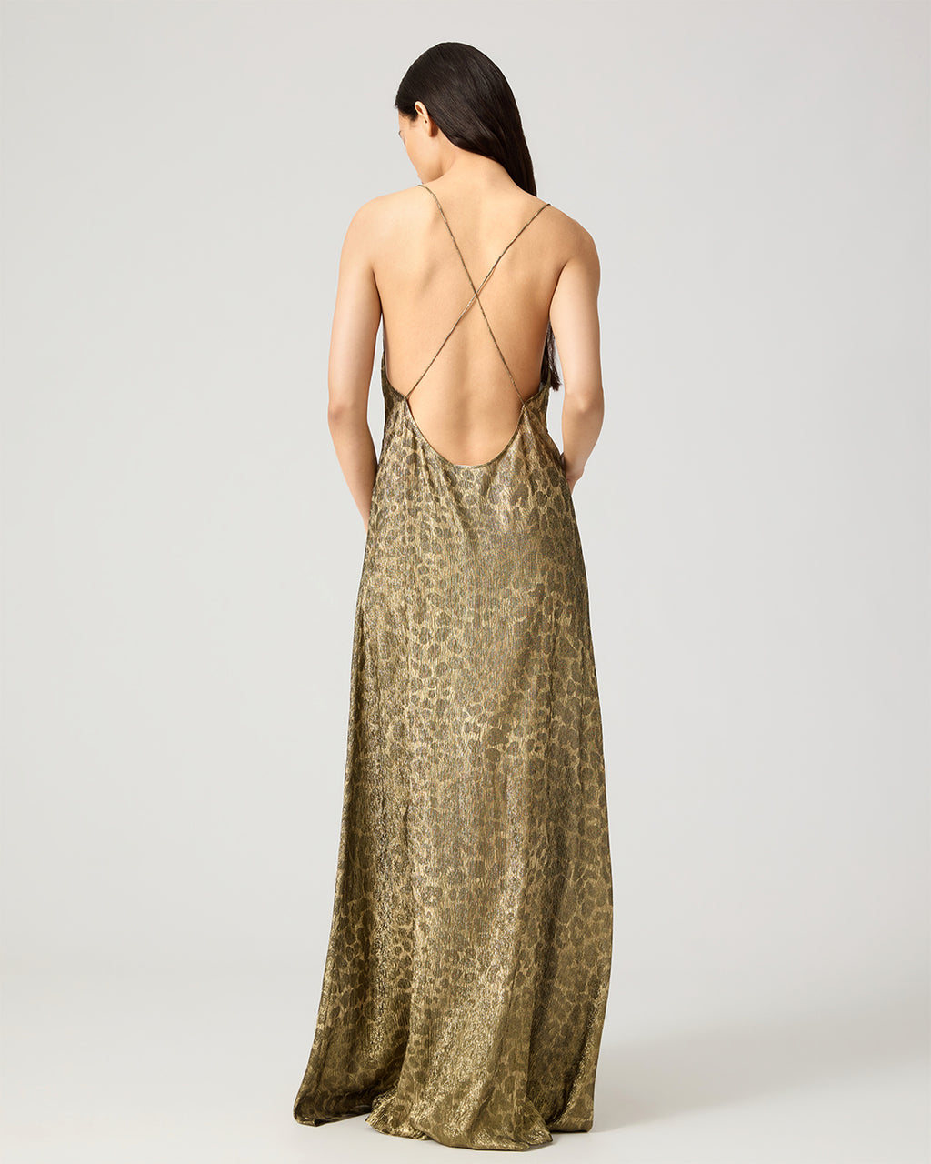 A woman shown from the back wearing a floor-length leopard-print slip dress with thin crisscross straps and an open back, photographed against a light gray studio background.