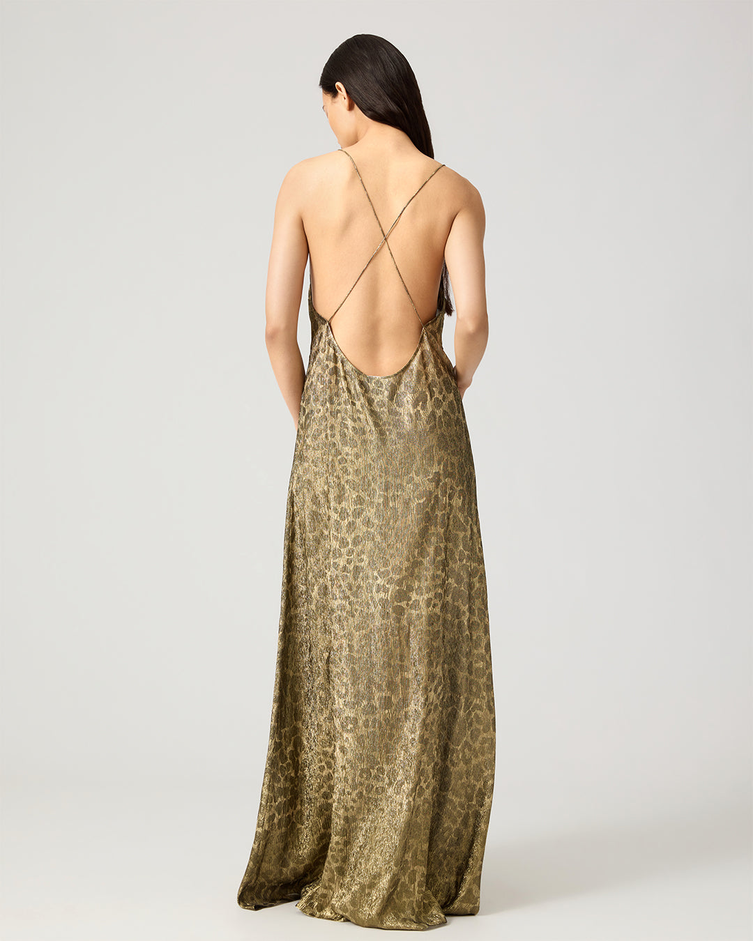 A woman shown from the back wearing a floor-length leopard-print slip dress with thin crisscross straps and an open back, photographed against a light gray studio background.