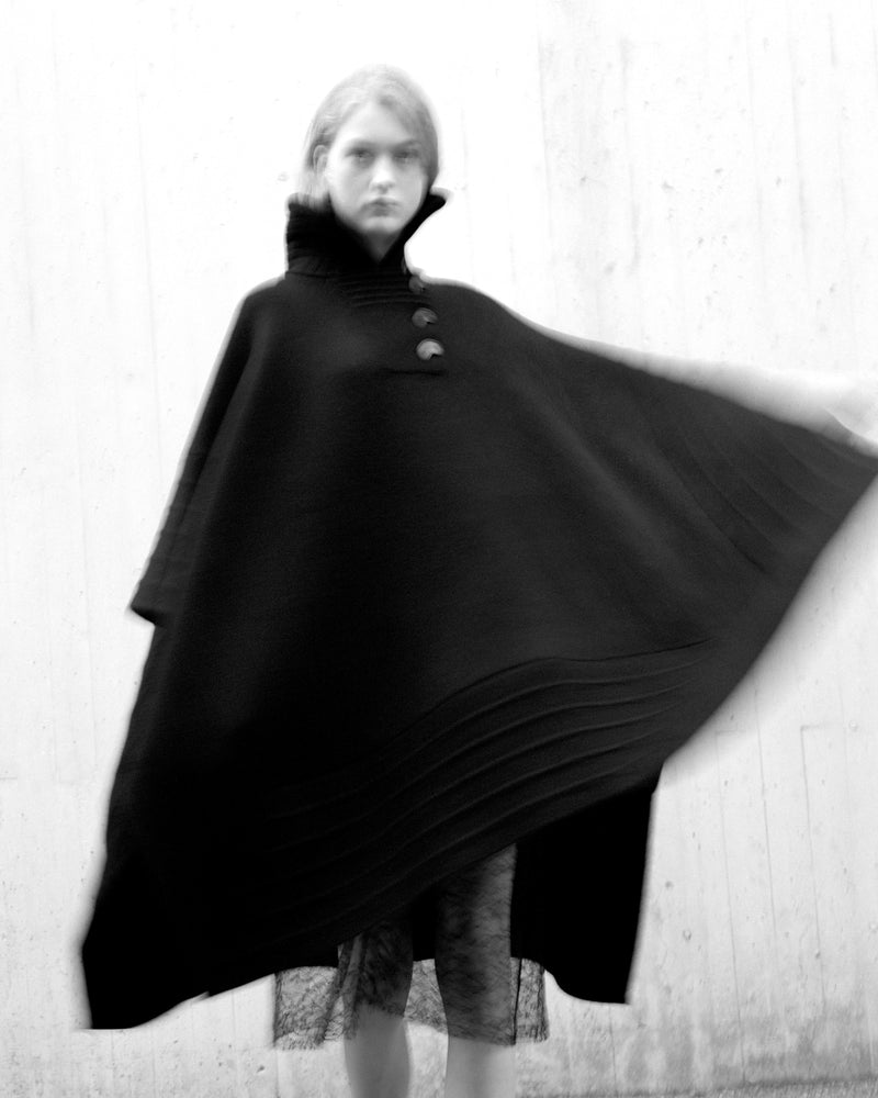 Black luxury women's cape with high funnel neck, three buttons, and pleated hem. Voluminous wool blend outerwear on model