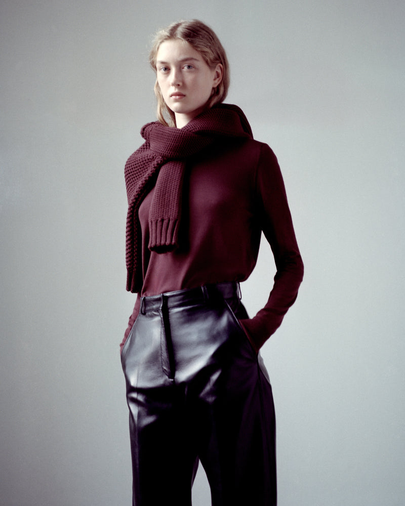 Woman in rich burgundy long-sleeve fine knit top, chunky knit scarf, black premium leather high-waisted pants